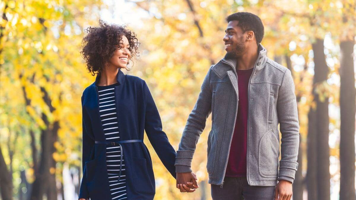 A couple holding hands and sharing a smile while walking among golden autumn trees