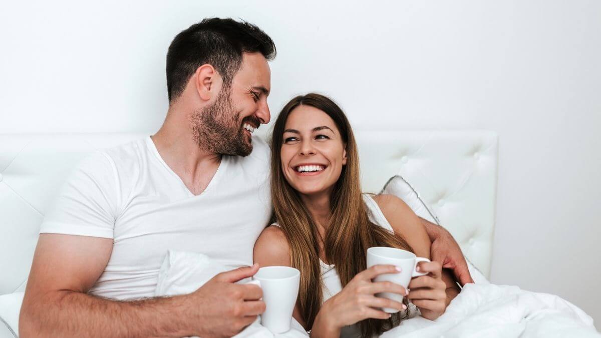 A couple laughing and holding coffee mugs while sitting up in bed