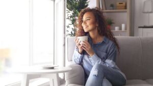 A woman sitting in an armchair holding a coffee cup and gazing out the window with a smile